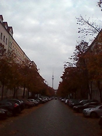 A street lined with parked cars and trees, leading to a tower in the distance on a cloudy day.