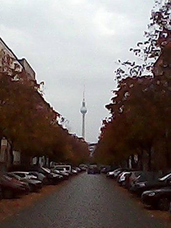 A street lined with parked cars and trees, leading to a tower in the distance on a cloudy day.