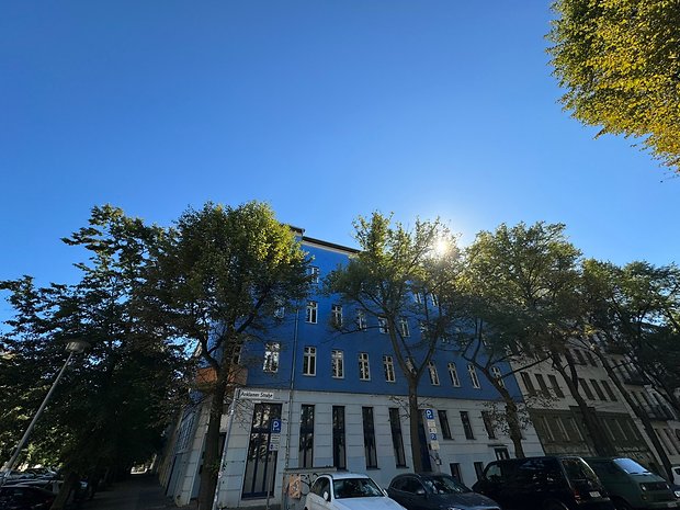 Photo prise avec l'iPhone 15 Pro Max de jour d'un bâtiment à l'angle d'une rue, aux murs bleu clair et avec des arbres au premier-plan