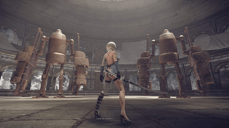 Eine Figur aus Nier: Automata steht mit einem Schwert in einem großen, runden Raum mit rostigen Roboterfiguren. Eine Figur aus Nier: Automata steht mit einem Schwert in einem großen, runden Raum mit rostigen Roboterfiguren.