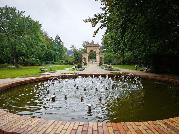 Une scène de parc avec une fontaine circulaire et une arche en arrière-plan, entourée de verdure et de bancs.