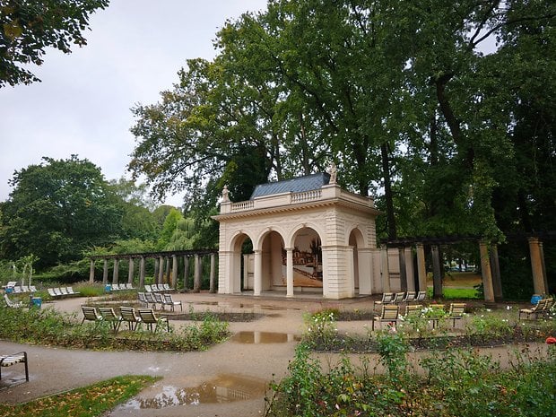 Un pavillon classique entouré d'arbres et de bancs vides dans un jardin après la pluie.