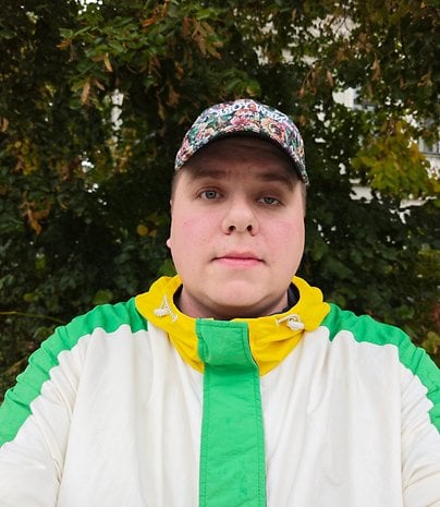 Une personne aux cheveux courts portant une veste colorée et une casquette se tient devant un feuillage vert.