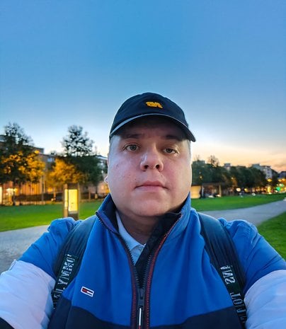 Une personne prend un selfie en plein air au coucher du soleil dans un parc, portant une veste bleue et une casquette.