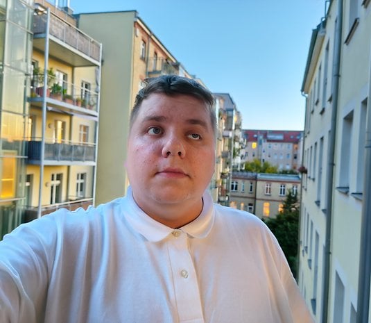 Un jeune homme en polo blanc se tient sur un balcon avec des bâtiments en arrière-plan.