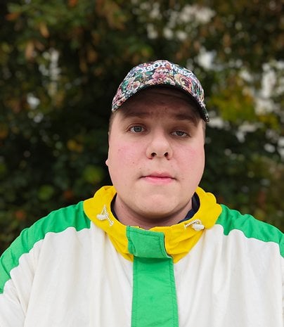 Une personne avec une casquette florale et une veste colorée se tient à l'extérieur devant de la verdure.