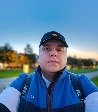 Un homme prenant un selfie en plein air, portant une veste bleue et une casquette noire, avec un flou d'arbres en arrière-plan.