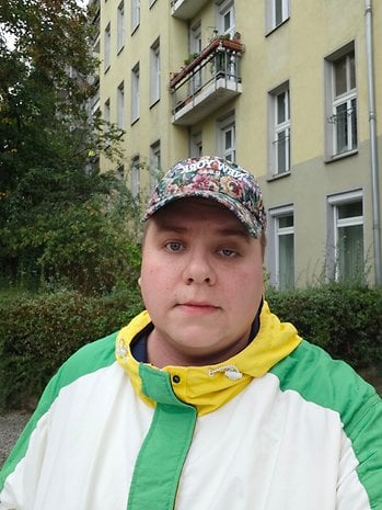 Une personne portant une veste colorée et une casquette se tient devant un bâtiment jaune avec des balcons.