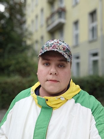 Une personne portant une veste colorée et une casquette à fleurs se tient devant un bâtiment jaune.