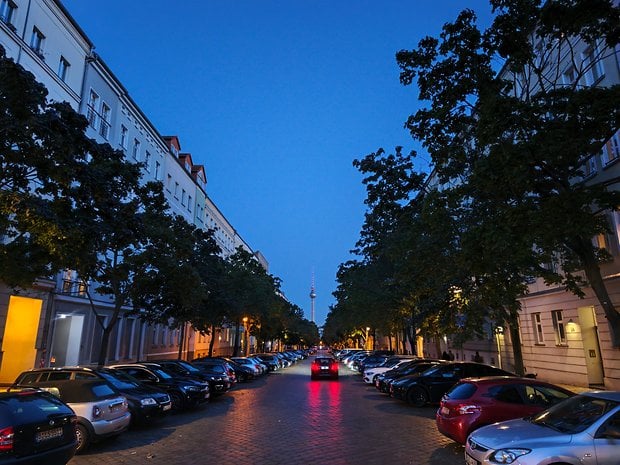 Une rue de la ville au crépuscule avec des voitures garées et des arbres des deux côtés, menant à une tour au loin.