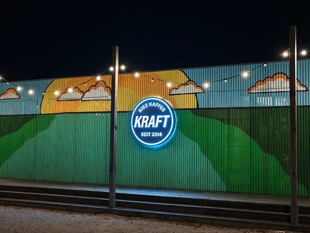 Une fresque murale colorée avec des collines verdoyantes et des nuages, et un néon bleu indiquant 'KRAFT seit 2014'.