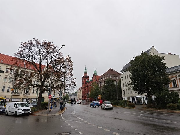 Vue de rue avec bâtiment rouge, arbres et voitures par une journée nuageuse.