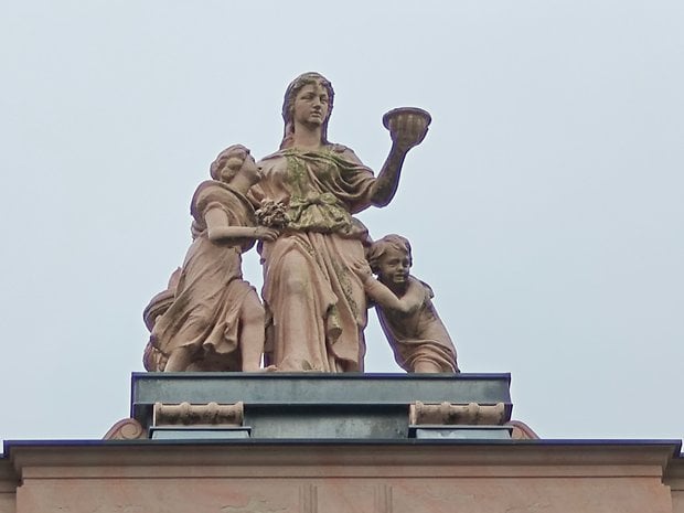 Statue d'une femme tenant un bol, accompagnée de deux enfants, contre un ciel nuageux.