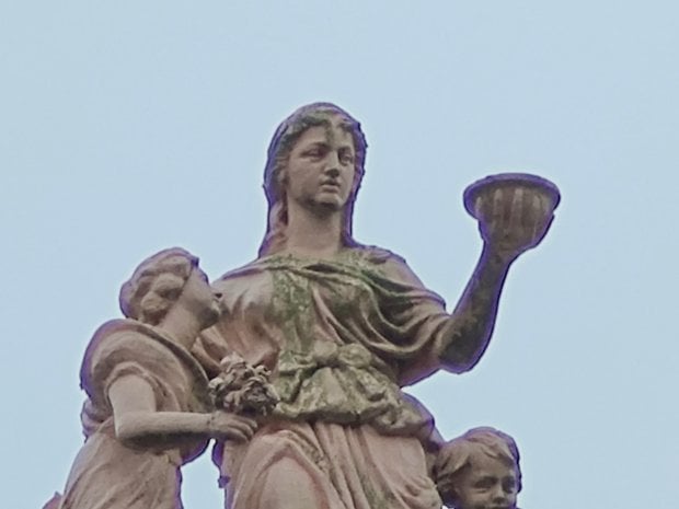 Une statue d'une femme tenant un bol, accompagnée de deux enfants, sur un ciel pâle.