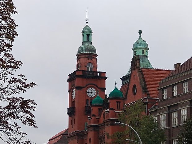 Bâtiment historique avec des dômes verts et une tour de l'horloge, entouré d'arbres par une journée nuageuse.