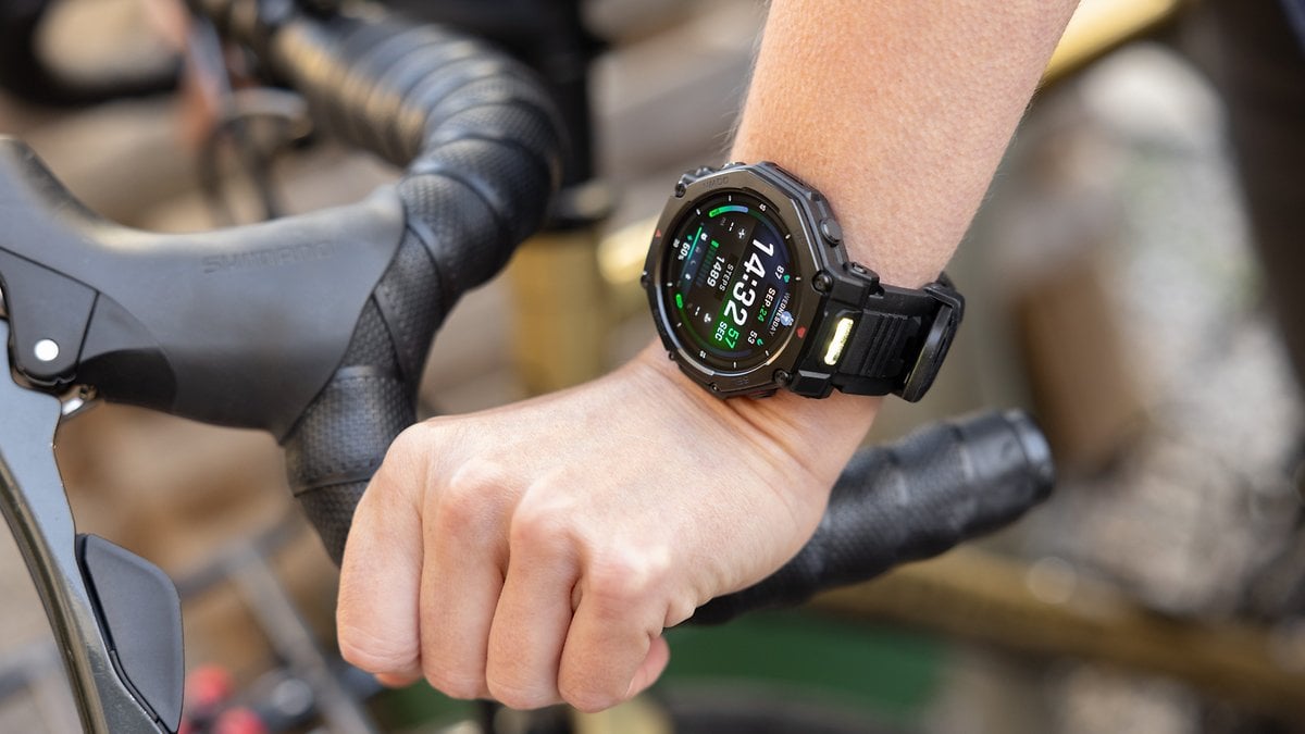 Una mano que lleva un smartwatch Amazfit T-Rex Pro 3 en el manillar de una bicicleta, mostrando la hora y datos de fitness.
