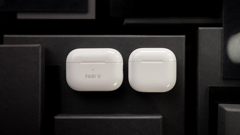 White Apple AirPods cases on a dark background, one with 'FABI' text. White Apple AirPods cases on a dark background, one with 'FABI' text.