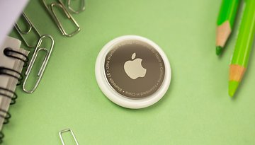 Ein Apple AirTag auf einer grünen Oberfläche, umgeben von Büroklammern und Buntstiften.