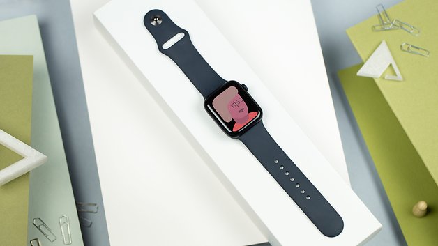 Um Apple Watch SE preto em uma base branca com um mostrador rosa mostrando a hora 11:15.