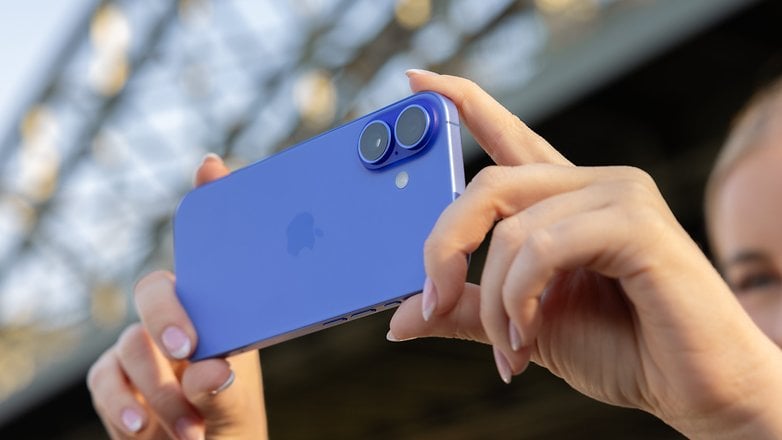 Eine Person hält ein blaues iPhone 16 mit Dual-Kameras in einer sonnigen Außenumgebung.