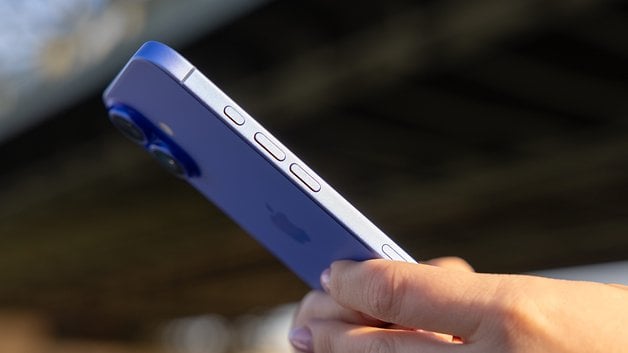 A person holding a blue iPhone 16, showing its rear and buttons against a blurred background.