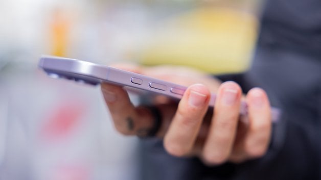 A person holding a purple iPhone 17, showing the side buttons and a blurred background.