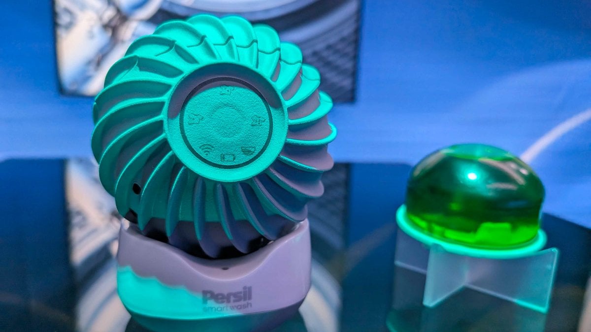 Un appareil vert en forme de ventilateur étiqueté 'Persil smartwash' à côté d'une capsule gélifiée verte sur une surface réfléchissante.