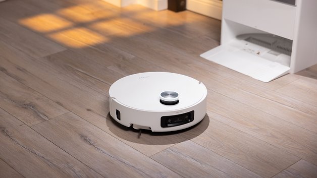 Ein weißer Roboterstaubsauger auf einem Holzboden, mit einer Ladestation im Hintergrund.