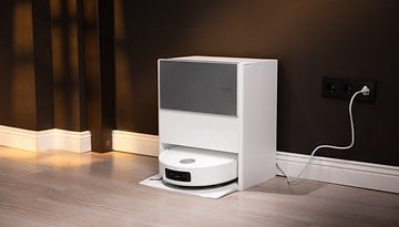 Ein weißer automatischer Staubsauger, angedockt an einer dunklen Wand, mit einem angeschlossenen Stromkabel.