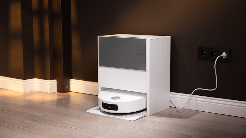 Ein weißer automatischer Staubsauger, angedockt an einer dunklen Wand, mit einem angeschlossenen Stromkabel. Ein weißer automatischer Staubsauger, angedockt an einer dunklen Wand, mit einem angeschlossenen Stromkabel.