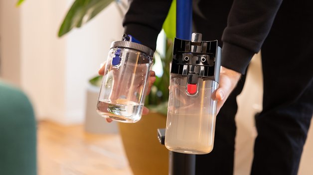 Eine Person hält zwei Behälter: einen klaren mit Wasser und einen trüben, die an einem Dyson-Staubsauger befestigt sind.