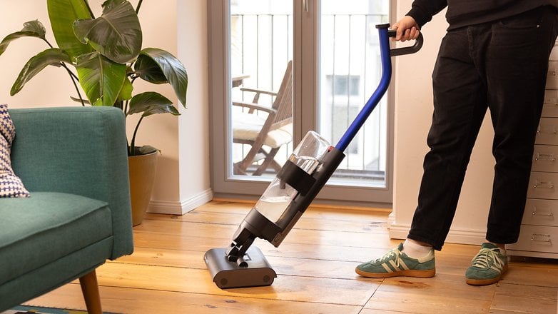 A person using a Dyson vacuum cleaner in a stylish living room with plants and a green couch.