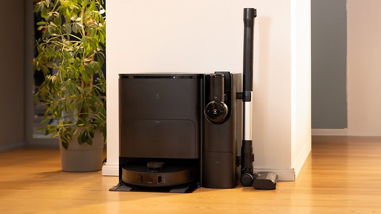 A black Ecovacs Deebot T30s Combo robot vacuum and its charging dock, placed next to a potted plant.