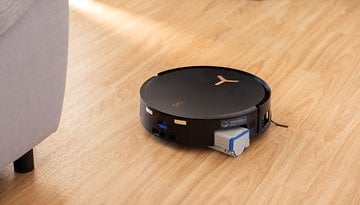 A black Ecovacs Deebot X8 Pro Omni robot vacuum cleaner on a wooden floor near a couch.