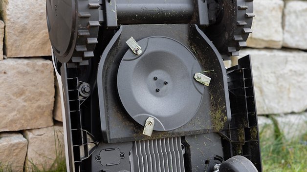 Gros plan du dessous d'une tondeuse à gazon robotisée avec des lames et des résidus d'herbe.