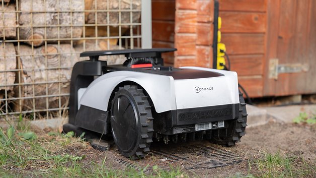 Une tondeuse robot Ecovacs garée sur l'herbe à côté d'une structure en bois et d'un empilement de bois de chauffage.