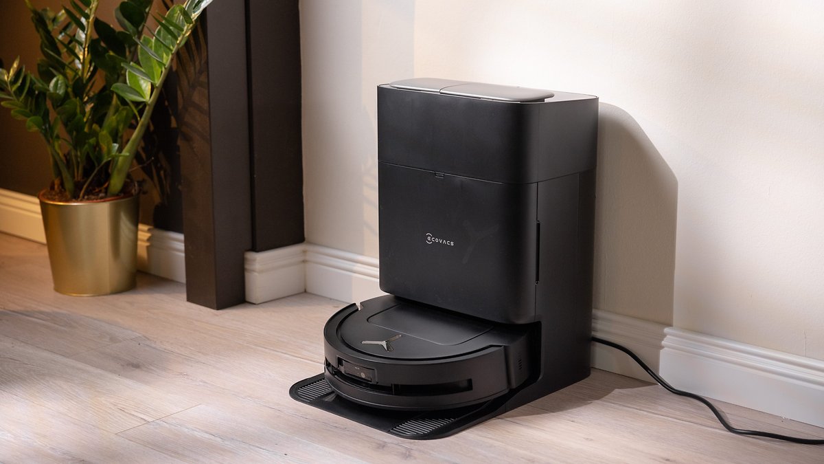 Un aspirateur robot Ecovacs T50 Omni noir à côté d'une plante contre un mur.