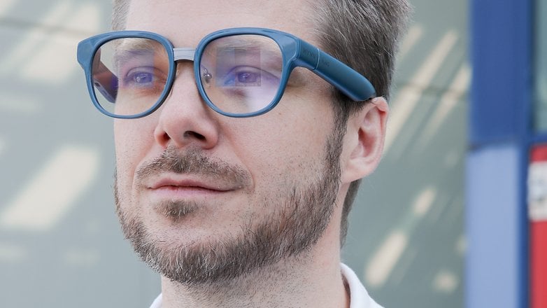 A man with a light beard wearing blue glasses, looking thoughtfully into the distance.