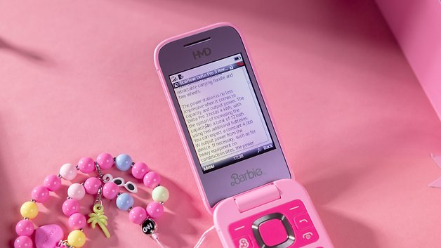 A pink flip phone with the 'Barbie' logo and text displayed on the screen, next to colorful beads.