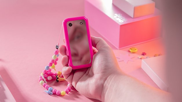 A pink Barbie phone with a mirror, held by one hand, on a pink background with decorative objects.