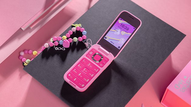 A pink flip phone on a black surface, displaying the time and surrounded by colorful jewels.