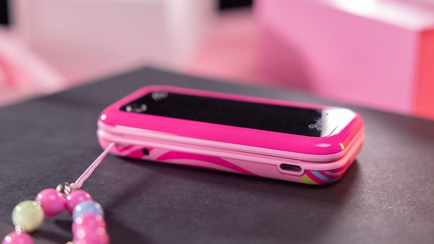 A pink flip phone with a black screen and colorful patterns, sitting on a dark surface.