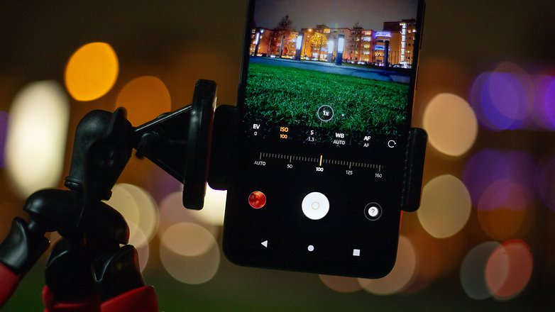 A smartphone mounted on a tripod displaying night photography settings with blurred colorful lights in the background.