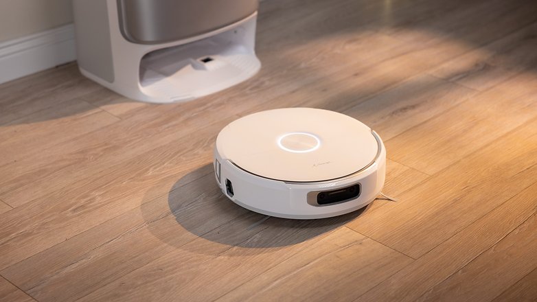 A white robotic vacuum cleaner on wooden floor, with a silver device in the background. A white robotic vacuum cleaner on wooden floor, with a silver device in the background.