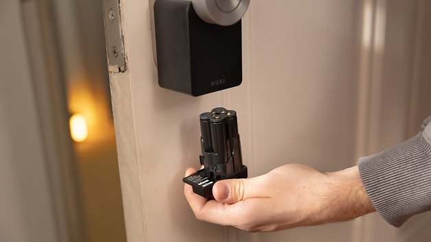 Eine Person hält das Nuki Smart Lock Pro, während sie neben einer Tür steht, an der das Schloss installiert ist.