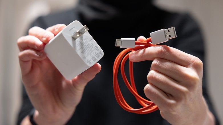 A person holding a white charger and an orange USB-C cable for the OnePlus 13.