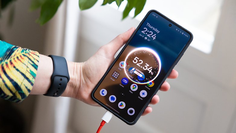 En person som håller en OnePlus 13R-smarttelefon som visar tid och batteristatus, ansluten till en laddare.