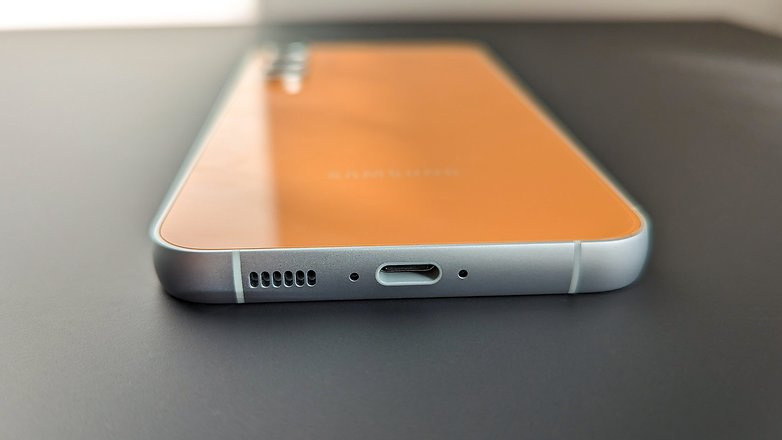 The S23 FE in the color tangerine lies on the table with the display facing downwards, view of the USB-C port from below.