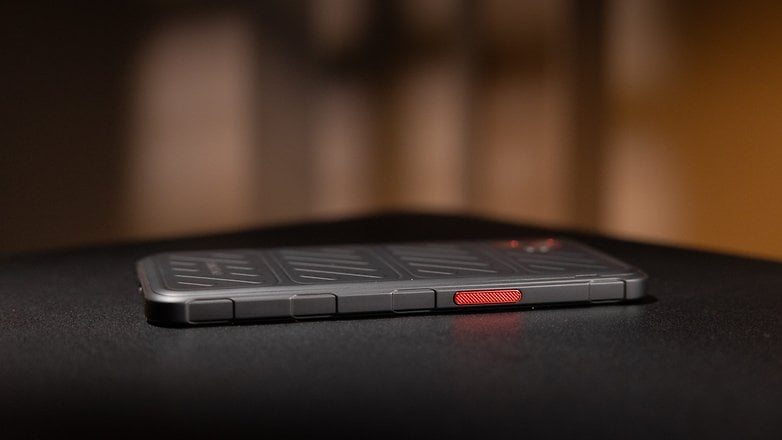 Samsung Galaxy XCover 7 Pro smartphone on a dark surface, showing a textured back and a red button. Samsung Galaxy XCover 7 Pro smartphone on a dark surface, showing a textured back and a red button.
