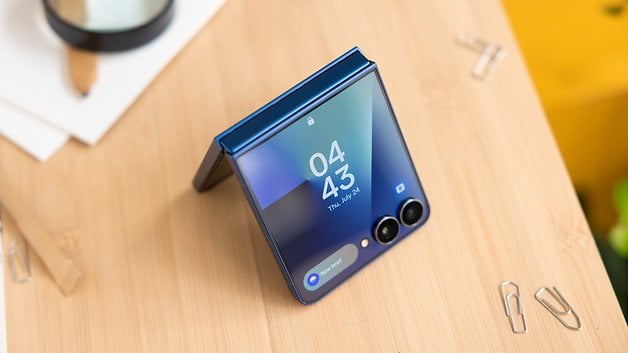Ein blauer Samsung Galaxy Z Flip7-Smartphone auf einer Holzoberfläche, die die Uhrzeit 04:43 anzeigt.
